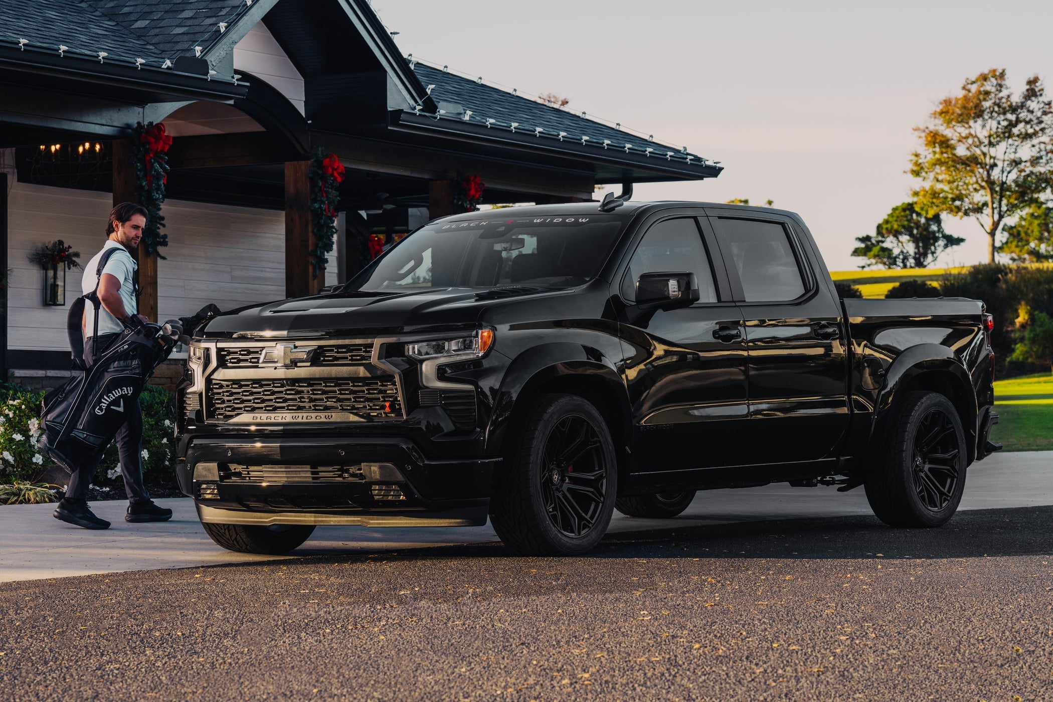A Black Widow lowered Silverado 1500 parked in front of a country club with a man with golf clubs walking by, looking at the truck in admiration.